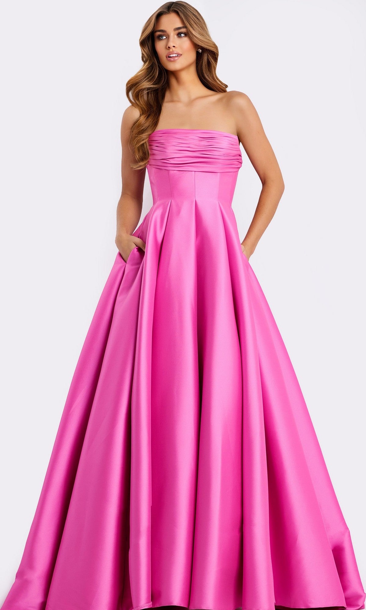 A woman stands against a plain background, wearing the Jovani 47570 Strapless Long Pleated Prom Dress featuring a fitted bodice and flowing A-line skirt.